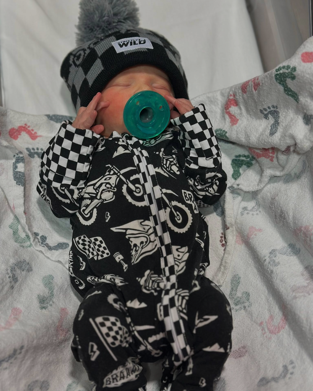 Baby in a black and white checkered outfit with a green pacifier, lying on a patterned blanket.