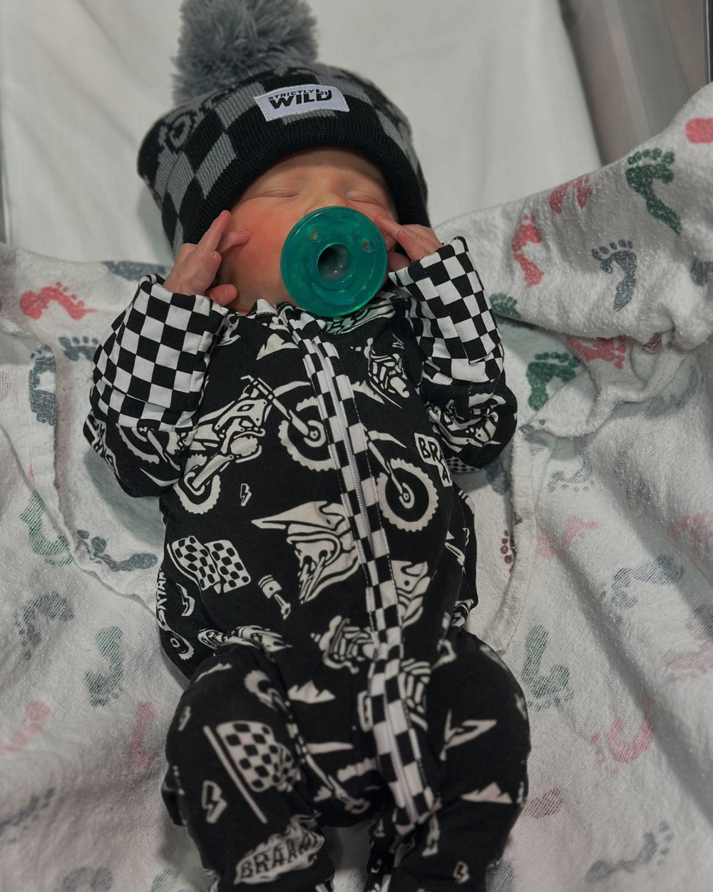 Baby in a black and white checkered outfit with a green pacifier, lying on a patterned blanket.