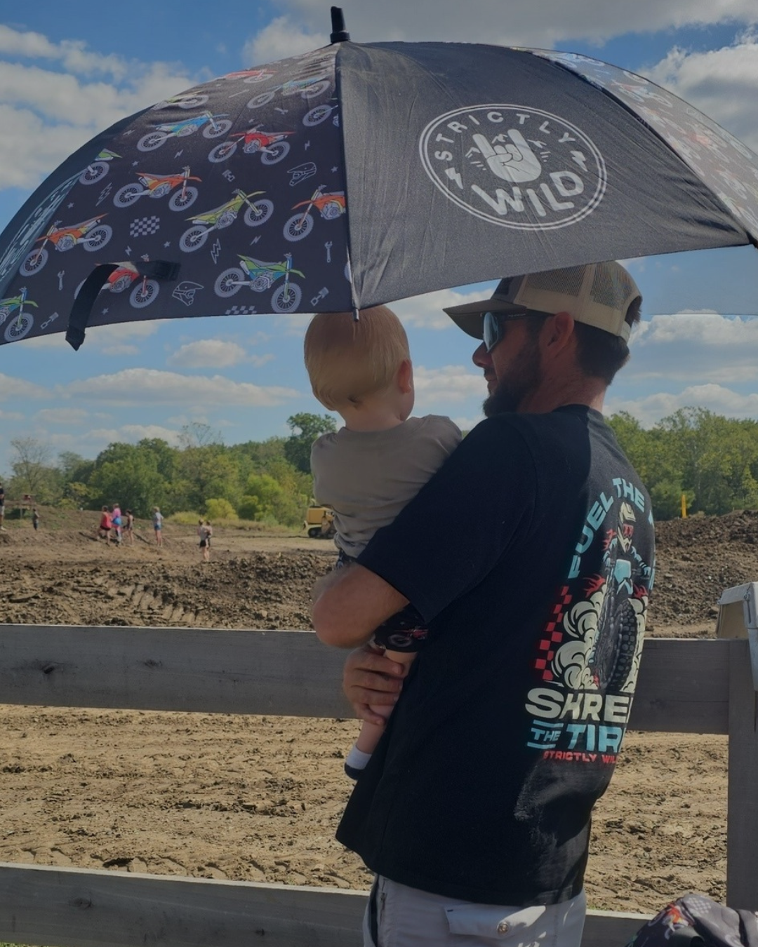 Man holding a child under an umbrella with a dirt field and people in the background