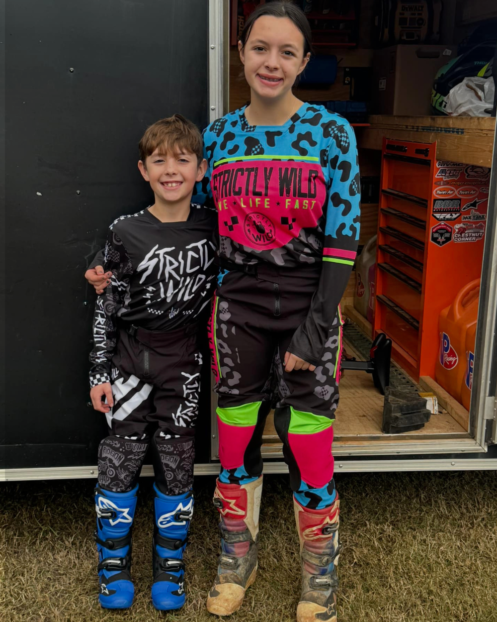 Two children wearing matching motocross gear standing in front of a trailer.