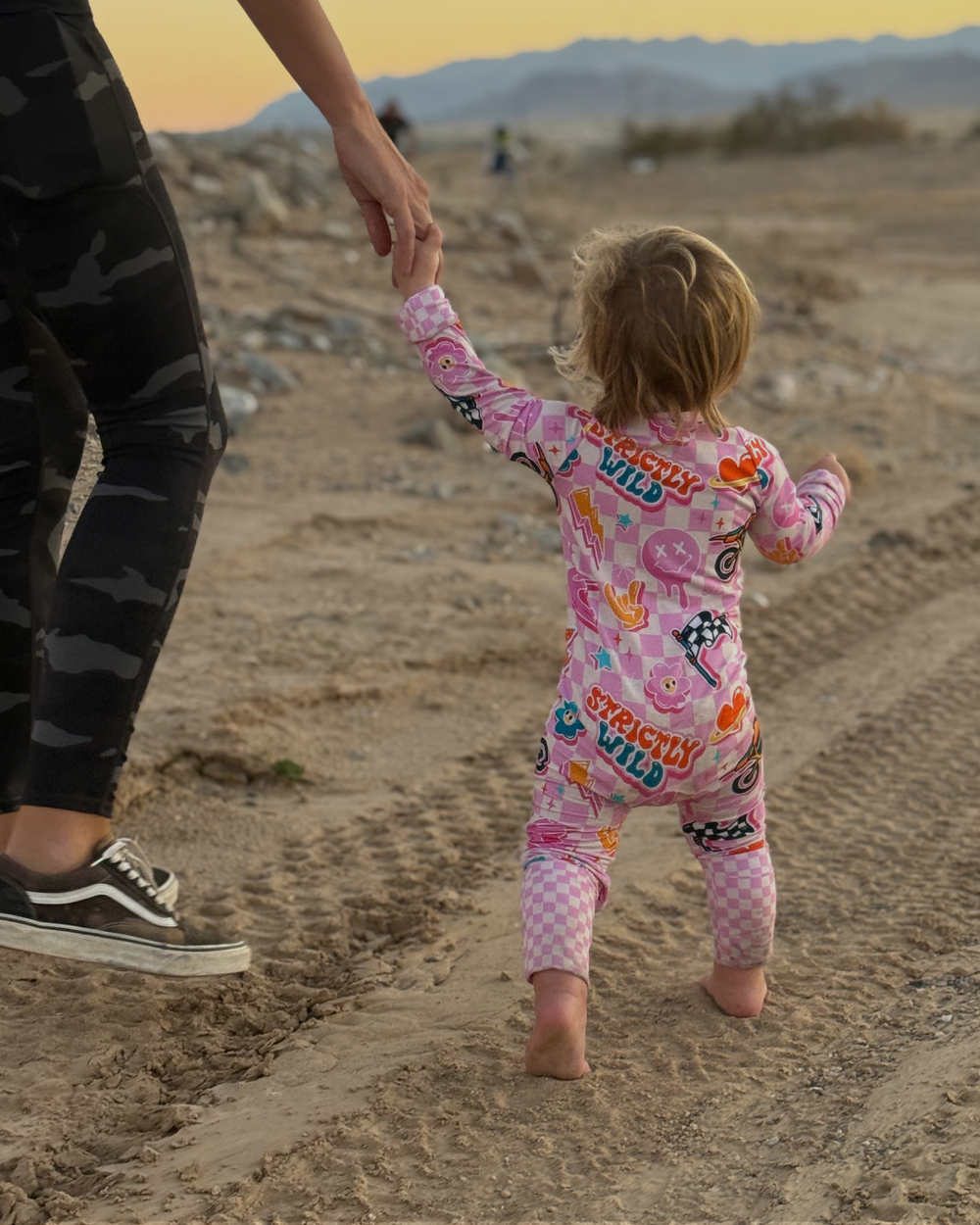 Child in a pink outfit walking on a desert path with an adult's hand, against a sunset sky.