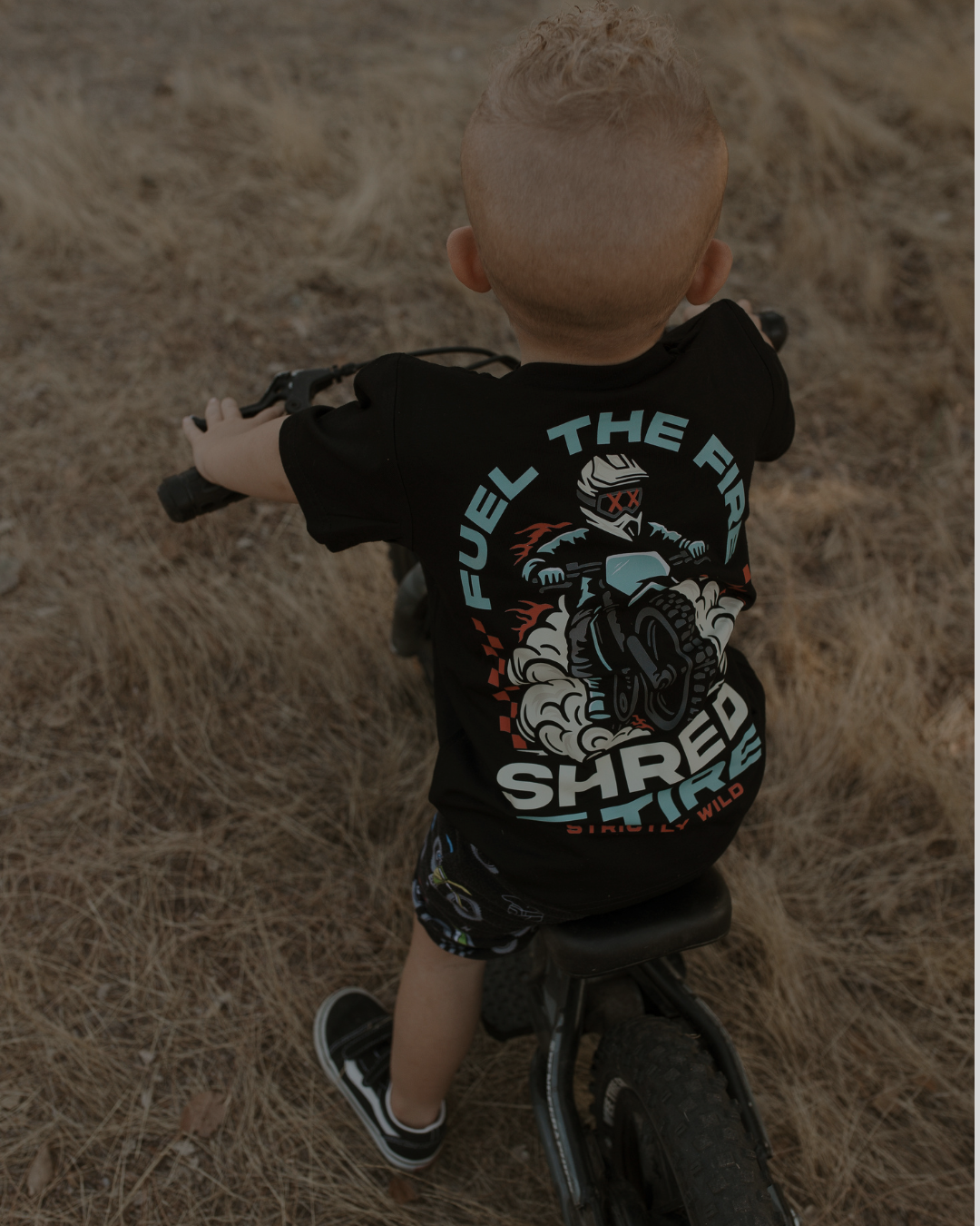 Child wearing a black t-shirt with graphic design and text, standing on a bike in a natural setting.