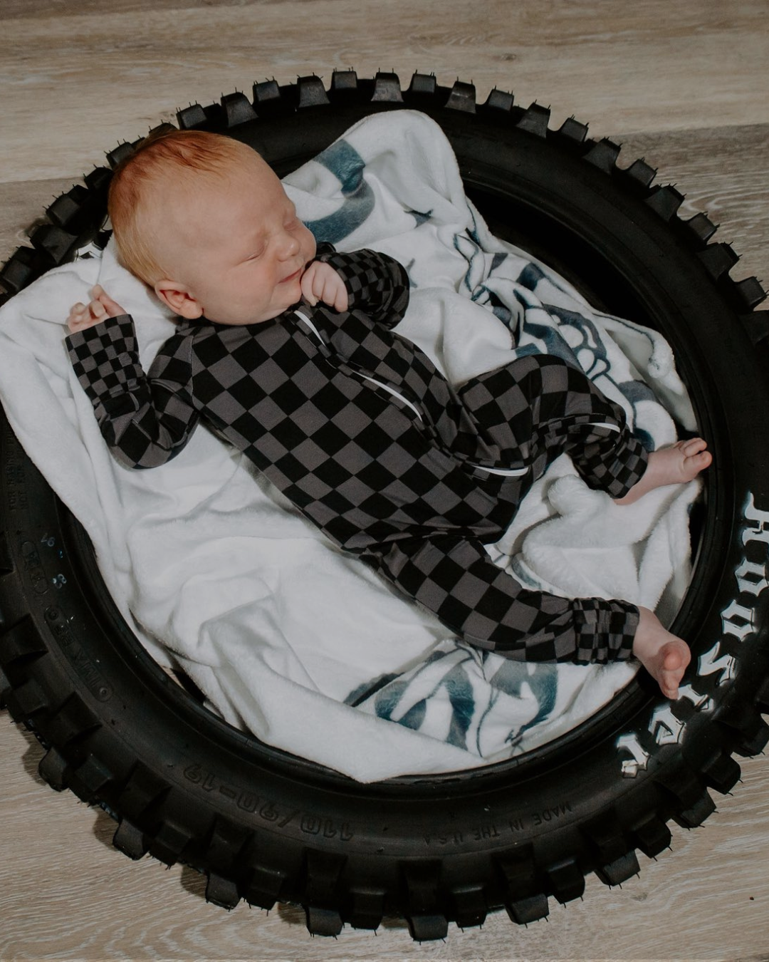 Newborn baby in a black and white checkered outfit lying on a white blanket inside a circular black crib.