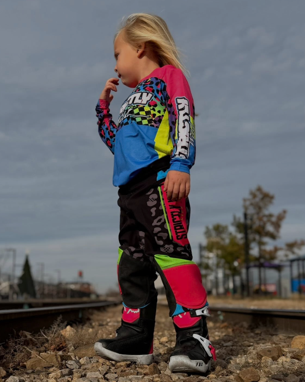 Child wearing a colorful motocross outfit standing on train tracks with a cloudy sky.