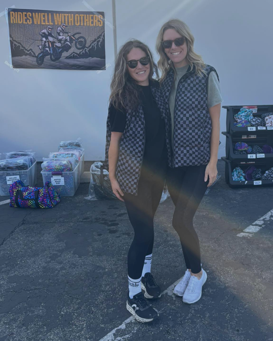 Two women standing together in a parking lot with various items and a poster in the background.