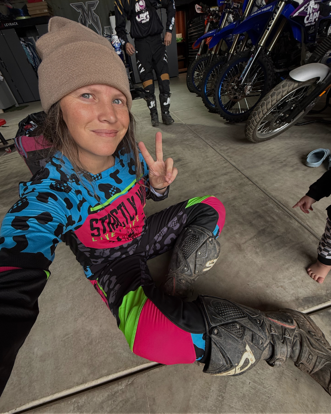 Person in motocross gear making a peace sign in a garage with motorcycles in the background