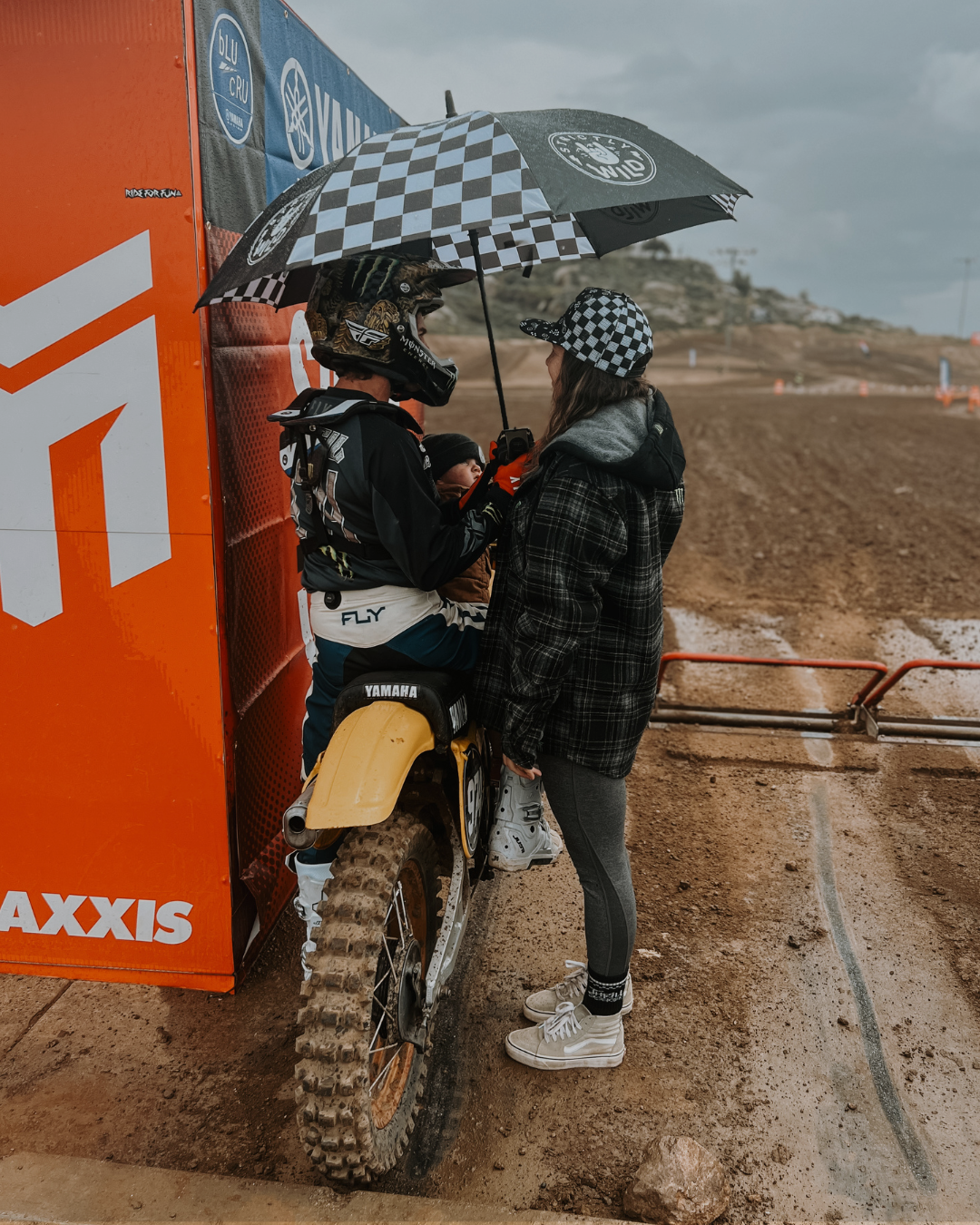 Person on a dirt bike with another person holding an umbrella under a checkered pattern, near a large orange 'AXXIS' sign.